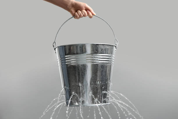Woman holding leaky bucket with water on light grey background, closeup Woman holding leaky bucket with water on light grey background, closeup leaking stock pictures, royalty-free photos & images