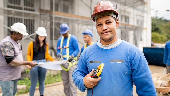 Portrait of a construction electrician holding a multimeter tool standing on a building site with people going over blueprints in the background