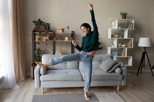 Full length overjoyed woman in casual clothes dancing in cozy living room, celebrating success, moving to popular music, moving into first new apartment, attractive excited young female having fun
