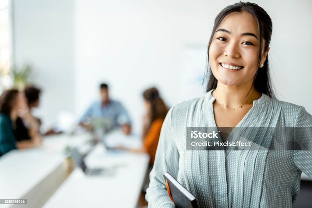 Asian Businesswoman Standing Smiling at the Camera Confident young Asian businesswoman standing smiling at the camera in a boardroom with colleagues in the background Employee Stock Photo