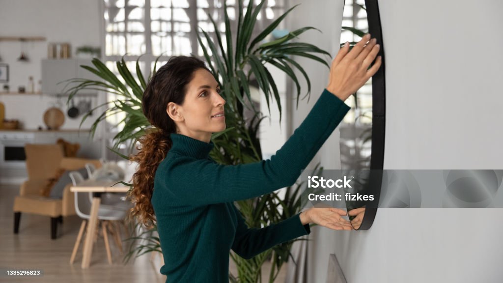Close up smiling woman hanging or fixing mirror, decorating apartment Close up smiling woman hanging or fixing mirror on wall, happy young female tenant renter decorating first own apartment, modern cozy living room, rent or mortgage concept, moving or relocation Mirror - Object Stock Photo