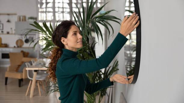 Close up smiling woman hanging or fixing mirror, decorating apartment stock photo