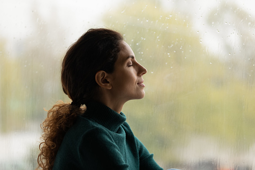 Profile view close up satisfied mindful woman with closed eyes dreaming near rainy window, meditating or daydreaming, visualizing good future, new opportunities, breathing deep, enjoying weather