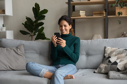 Smiling young woman relaxing on couch with smartphone, browsing mobile device apps, looking at phone screen, chatting or shopping online, enjoying leisure time with gadget, watching funny video