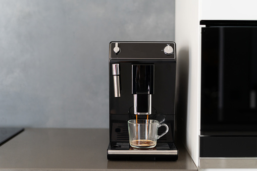Coffee making machine pouring espresso into glass, getting ready and energized before going to work, kitchen counter with home appliances, cupboard with integrated technology