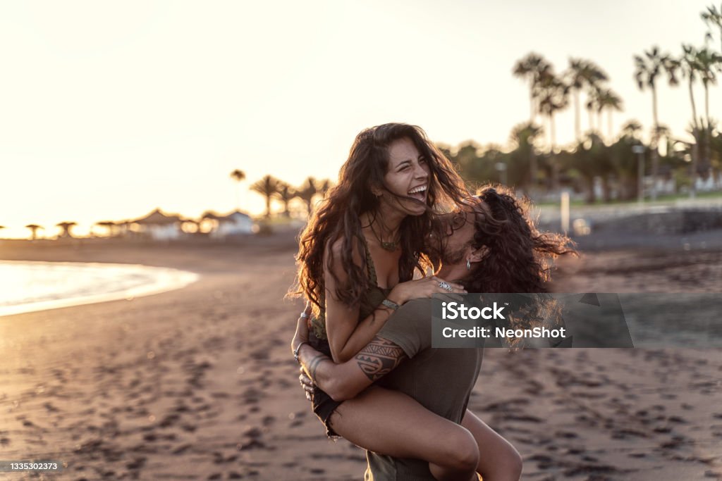 Happy beautfiul italian couple having fun together on the beach, laughing. Sunset time. Happy beautfiul italian couple having fun together on the beach, laughing. Sunset time. Summer island vibes. Real people emotions and lifestyle. Couple - Relationship Stock Photo