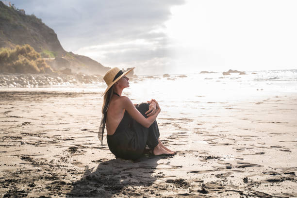 mujer tranquila sentada sola en la playa durante la puesta del sol, respirando, meditando. - one person beautiful barefoot beach fotografías e imágenes de stock