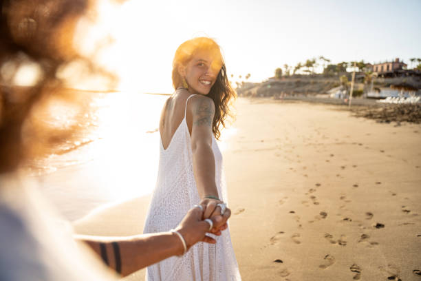 urocza uśmiechnięta piękna kobieta trzymająca za rękę swojego chłopaka, patrząc na niego, spacerując po plaży - rugged coastline zdjęcia i obrazy z banku zdjęć