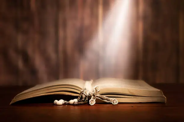 Dramatic image showing a bright light beam shining on an old bible with a rosary laying in front of it.