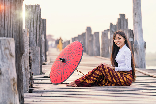A young Burmese woman wearing traditional clothes with red umbrella at  U Bein teak bridge on Taungthaman lake in Mandalay Division. Myanmar.