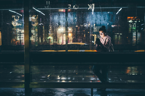 Young woman waiting for public transport inside modern transparent shelter at the night.