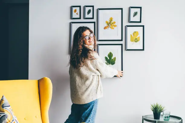 Photo of Skilled woman designer with long loose curly hair holds wooden frame with dried leaf applique and hangs on decorated wall in new apartment backside view.