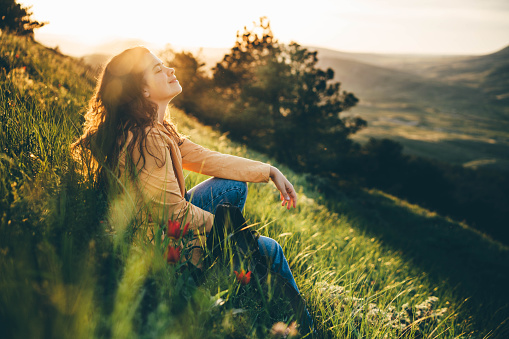 La joven viajera con el pelo rizado suelto largo se sienta en el prado verde de la hierba con las flores y los tipos en el teléfono inteligente contra paisaje montañoso bajo luz del sol photo