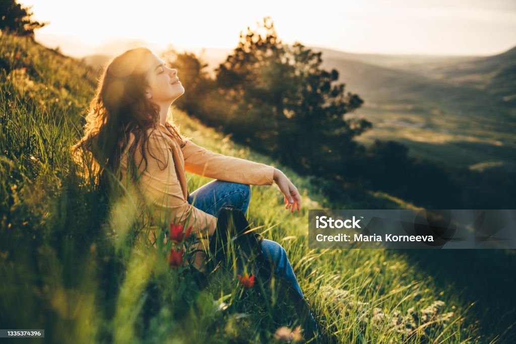 Young woman traveler with long loose curly hair sits on green grass meadow with flowers and types on smartphone against hilly landscape under sunlight Women Stock Photo