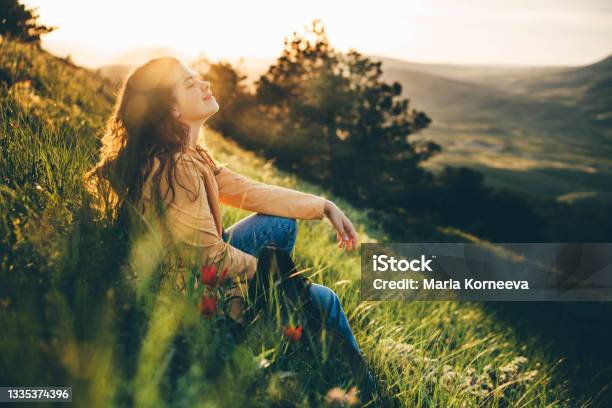 Young Woman Traveler With Long Loose Curly Hair Sits On Green Grass Meadow With Flowers And Types On Smartphone Against Hilly Landscape Under Sunlight Stock Photo - Download Image Now