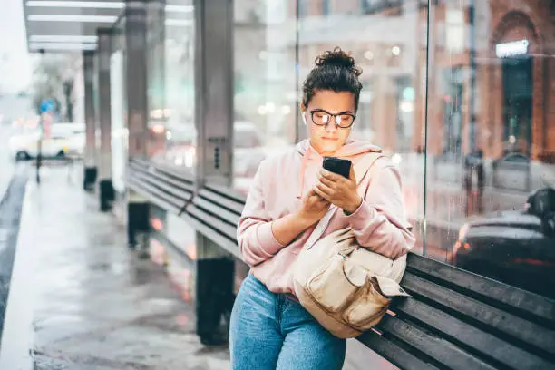 Photo of Woman waiting for public transport on bus stop looking to the smartphone, using application for searching traffic schedule.
