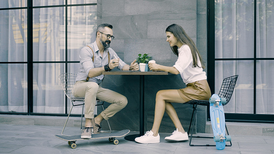 Middle age man sitting with his girlfriend at outdoor café with their skateboard using digital tablet