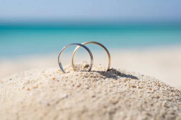 Pair of gold wedding ring bands jewelry in sand on tropical desert island beach during summer with blue ocean background