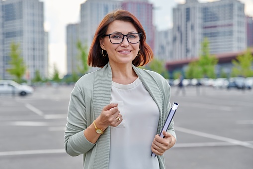Outdoor portrait of business woman 40s of age. Smiling female in glasses looking at camera with business notebook in hands, city buildings urban architecture background. Profession, middle aged people