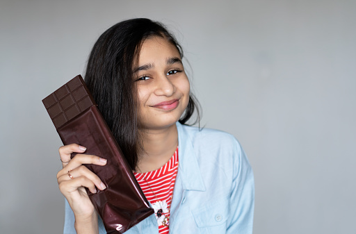 Happy Asian, Indian girl holding a big dark chocolate bar in hand. She is standing against gray background and looking at the camera with a smile.