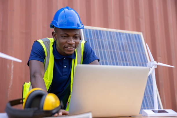 engenheiro afro-americano e eletricista caucasiano usando chapéu duro azul trabalhando em computador portátil na área de trabalho conceito de energia alternativa limpa e verde. - electricity control panel engineer electronics industry - fotografias e filmes do acervo