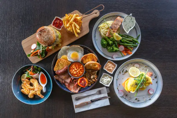 A top view of a table arranged with dishes of a burger menu with fries, English breakfast, salmon, cooked vegetables, fried prawns, cutlery and sauces.