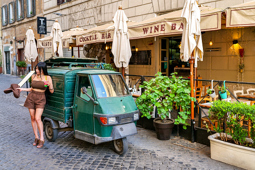 Rome, Italy, August 13 -- A young tourist poses for a photo near a characteristic Piaggio Ape along an alley in Campo de Fiori, in the historic and baroque heart of Rome. The Piaggio Ape is an Italian three-wheeled light commercial vehicle very popular in Italy, easy to drive and very economical, used in the historic centers of cities mainly for the delivery of food products. The district of Piazza Campo de Fiori is much loved and visited by tourists and young people for the presence of the famous fruit and vegetable market, but also for the presence of ancient churches and noble palaces, for the countless artistic and cultural treasures, for the many typical and trendy restaurants and the characteristic alleys to be discovered by walking freely, where you can experience the essence and soul of Roman life. In 1980 the historic center of Rome was declared a World Heritage Site by Unesco. Image in high definition format.