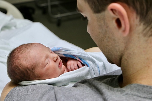 Contented emotion over the shoulder view of father lovingly holding newborn baby daughter in hospital.