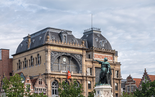 Gent, Flanders, Belgium - July 30, 2021: Jacob Van Artevelde statue in front of Socialist Bond Mouson historic building under blueish cloudscape. Green foliage and red flag.