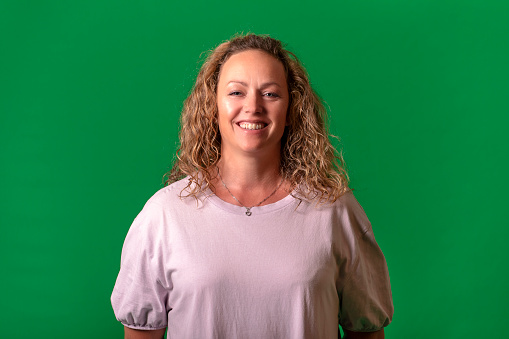Front portrait of mature blonde woman on green background. She has white blouse, happy smiling and looking at camera.