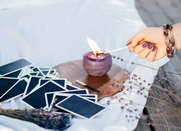 Woman's hand in bracelet with purple long nails lights candle with match, next to deck of cards and lavender flowers on white and wooden surface