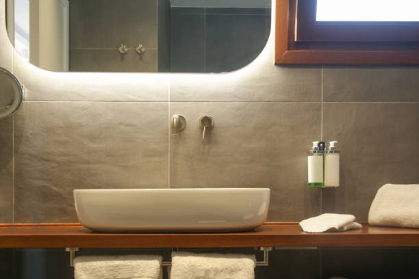 istock Modern simple bathroom with white bathroom sink standing on wooden shelf and backlit mirror 1335482881