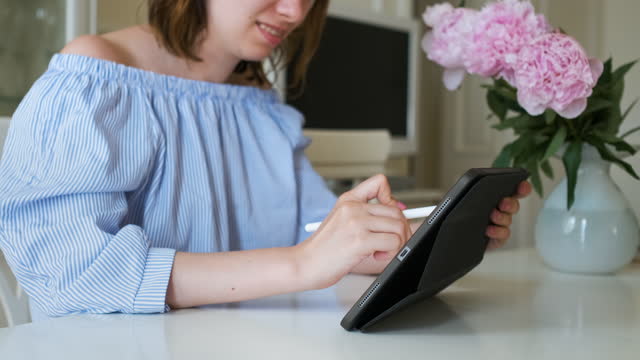 Young cheerful woman working on the tablet with pencil in modern interior with peonies on the background. Freelance or designer working at home.