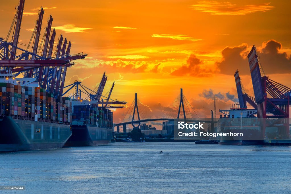 Harbor and a bridge in the morning Container ships, harbor cranes and a bridge in the far. Orange, morning sky in background. Small motorboat passing the dock. Port Of Hamburg Stock Photo