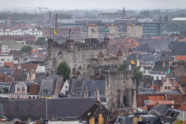Gent, Flanders, Belgium - July 30, 2021: Dark gray Gravensteen stone castle and its many flags surrounded by cityscape in rain. Hospital complex behind. Many facades and gables.
