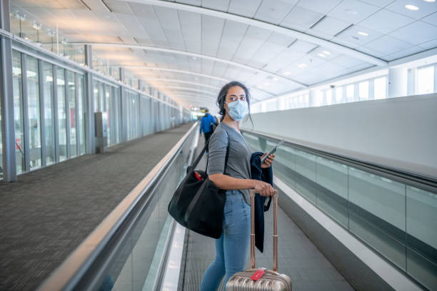 Solo young female traveler at the airport during the pandemic A young female traveler of middle eastern ethnicity is on a walking escalator at the airport. She has her carry on suit case and a bag on her. She is wearing a face mask to prevent the spread of germs. airports canada stock pictures, royalty-free photos & images