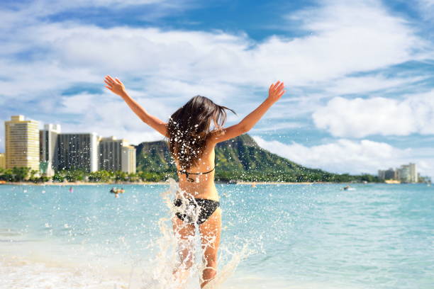 beach fun - femme heureuse sur hawaii waikiki vacances. jeune adulte méconnaissable de derrière sautant de joie dans les vagues d’eau, bras vers le haut avec la montagne de tête de diamant en arrière-plan, point de repère d’honolulu. - waikiki beach photos et images de collection