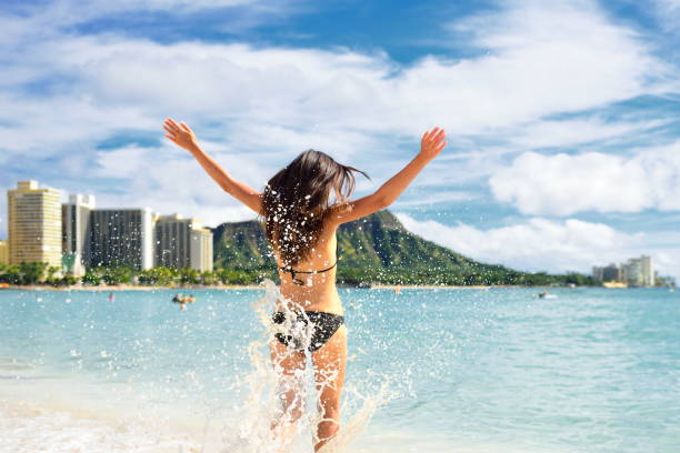 divertimento in spiaggia - donna felice durante le vacanze alle hawaii waikiki. giovane adulto irriconoscibile da dietro saltando di gioia nelle onde d'acqua, braccia su con la montagna testa di diamante sullo sfondo, punto di riferimento di honolulu. - waikiki beach foto e immagini stock