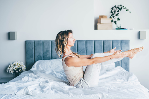 Lady stretches legs and changes yoga positions lying on bed among pillows against large plastic windows and curtains at bright light in morning