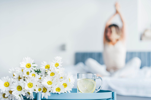 Morning concept - close up glass of water with lemon and flower in the foreground. And the woman is out of focus in the background.