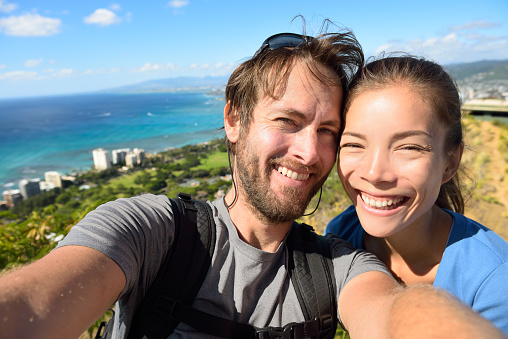 Selfie couple travel fun - Tourists on Hawaii taking selfie photo of Honolulu and Waikiki beach. Woman and man on hike visiting viewpoint lookout in Diamond Head State Monument and park, Oahu, USA.