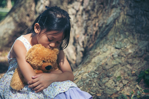 Sad girl hugging teddy bear sitting under tree sadness alone in green park. Lonely girl feeling sad unhappy sit outdoors hug best friend toy. Autism child play teddy bear best friend. Family violence