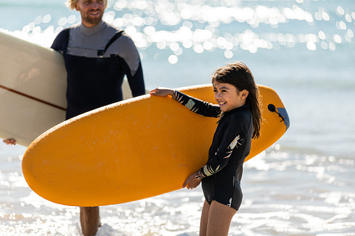 Mixed race girl carries her surfboard with her Dad nearby