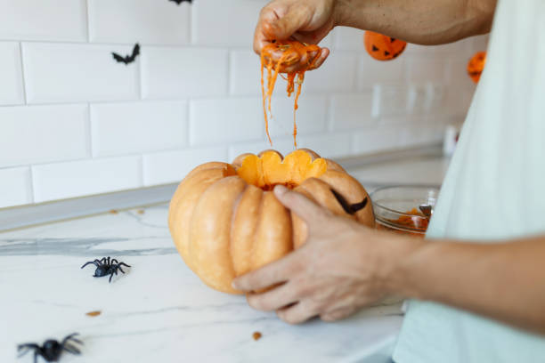 eine nahaufnahme der hand des mannes zieht samen und faseriges material aus einem kürbis, bevor sie für halloween schnitzt. jack-o-laterne. seitenansicht in der küche - photography close up side view halloween stock-fotos und bilder