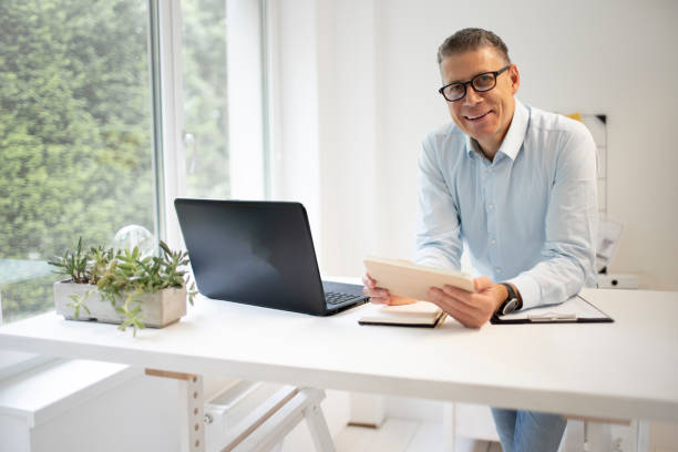 istock man with blue shirt and black glasses is standing behind standing table and is working with his tablet 1335540191