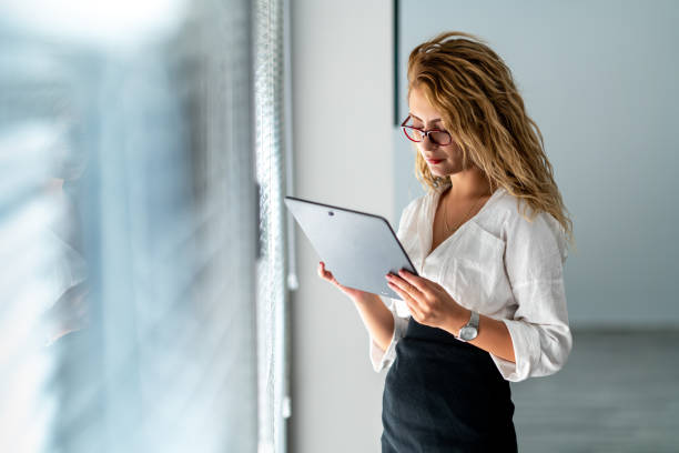 istock Young beautiful business woman is holding a mobile tablet on her hand, to check her work, while she is standing next to windows. 1335550654