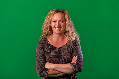 Front portrait of mature blonde woman on green background. She has dark grey blouse, happy smiling and looking at camera. Her hands are based on her chest.