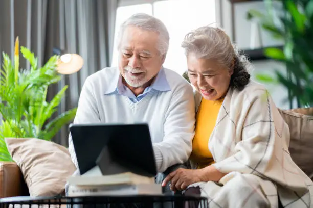 Photo of old asian senior couple retired age sitting on sofa couch video call to their family with tablet device social connect home isolation stay home state order concept,happiness senior laugh smile joyful