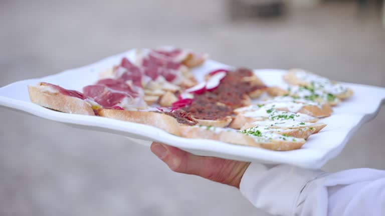 Close up of coming waiter holding plate with assortment of snacks and canapes, tasty appetizing bread slices with Italian prosciutto, vegetables and sauce toppings for luxury wedding party outdoors. Catering food service for rich people