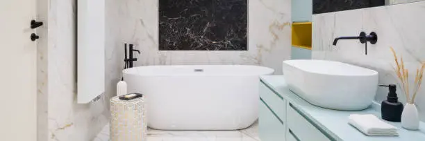 Panorama of elegant bathroom with black and white marble tiles on floor and walls, big freestanding bathtub and blue chest of drawers under washbasin